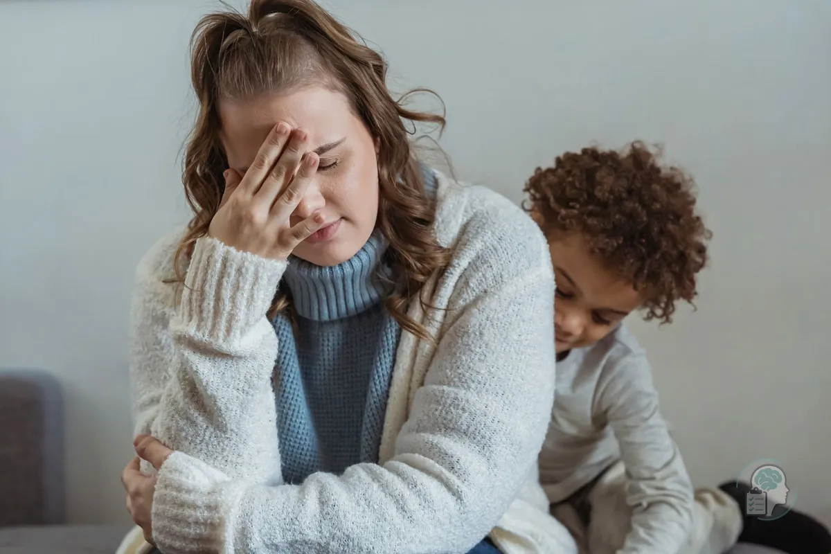 Madre agotada que se tapa la cara con la mano, sentada en el sofá mientras su hijo pequeño juega a su lado, un ejemplo de agotamiento emocional en la crianza