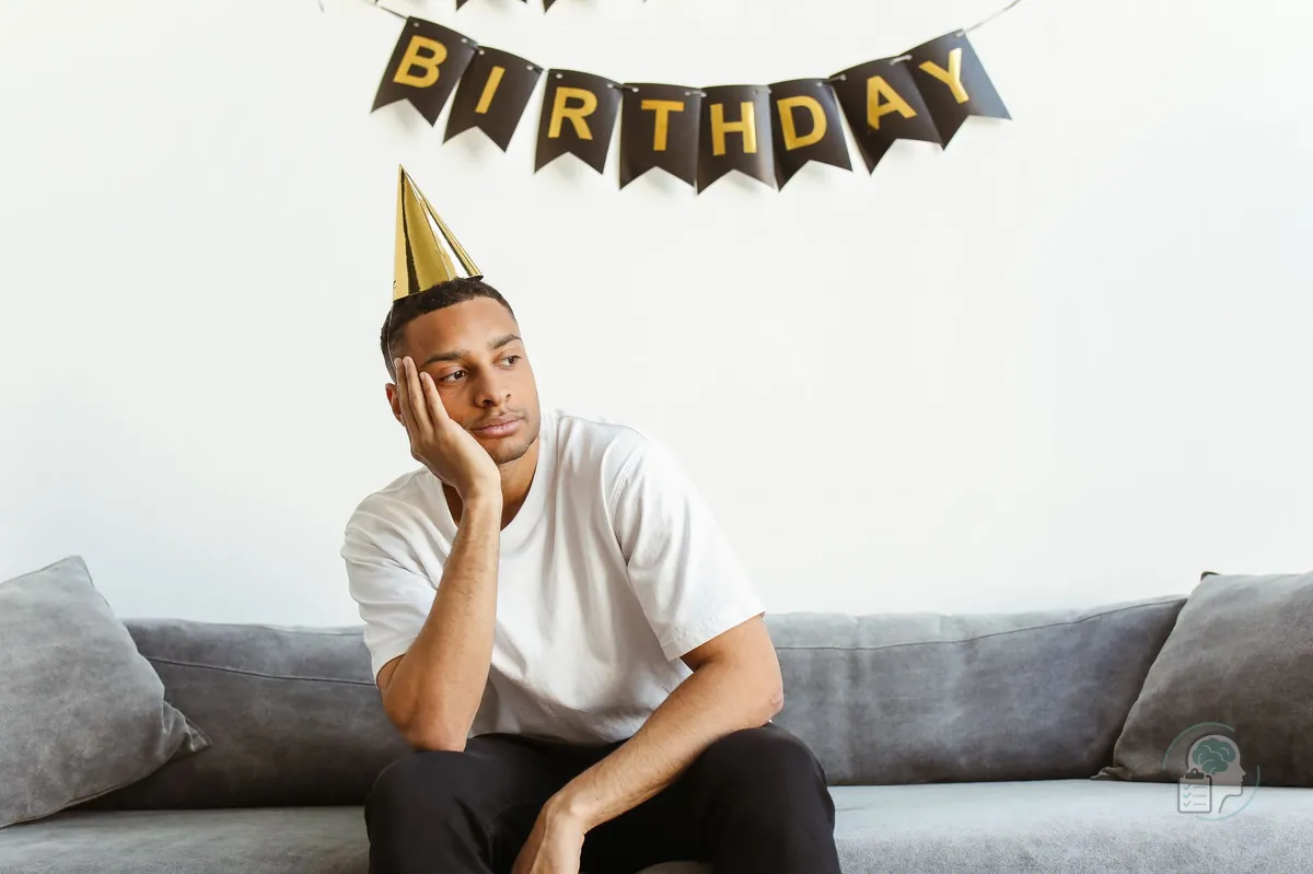 Hombre con gorro de fiesta sentado en el sofá bajo una guirnalda de cumpleaños, con gesto de aburrimiento, una imagen de la anhedonia