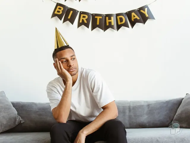 Hombre con gorro de fiesta sentado en el sofá bajo una guirnalda de cumpleaños, con gesto de aburrimiento, una imagen de la anhedonia