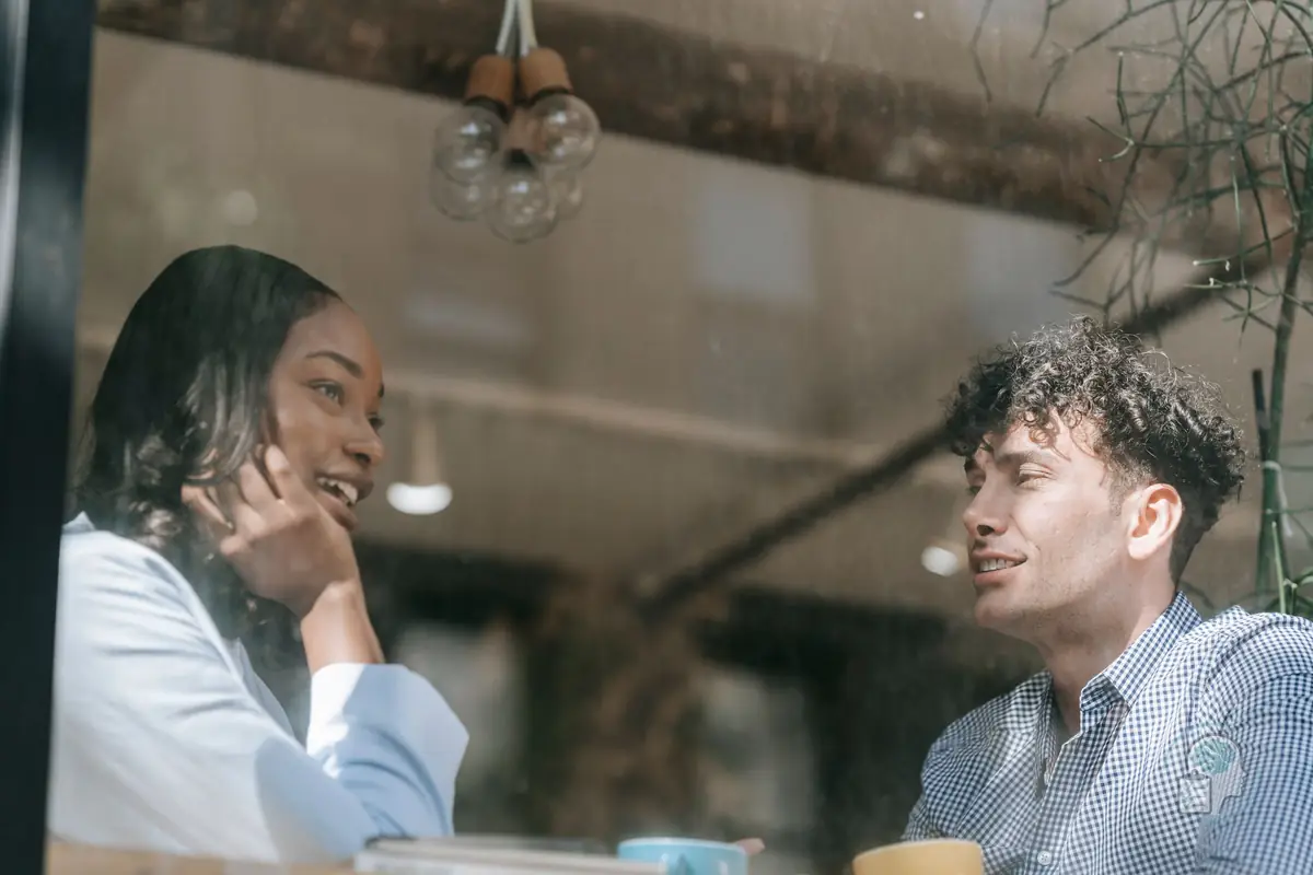 Mujer que escucha sonriente a un amigo en la mesa de una cafetería, con la atención puesta en la conversación