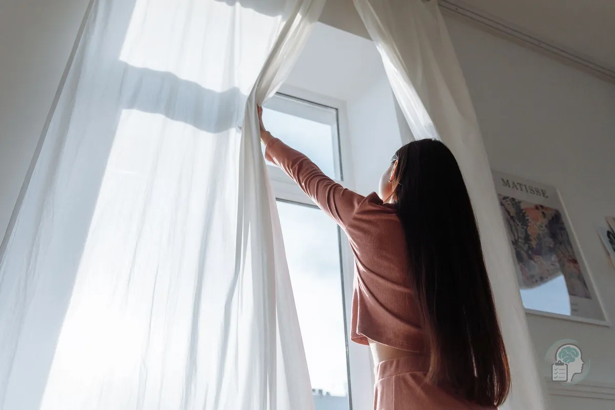 Mujer que abre las cortinas para dejar entrar la luz de la mañana, un gesto sencillo que ayuda en la depresión estacional