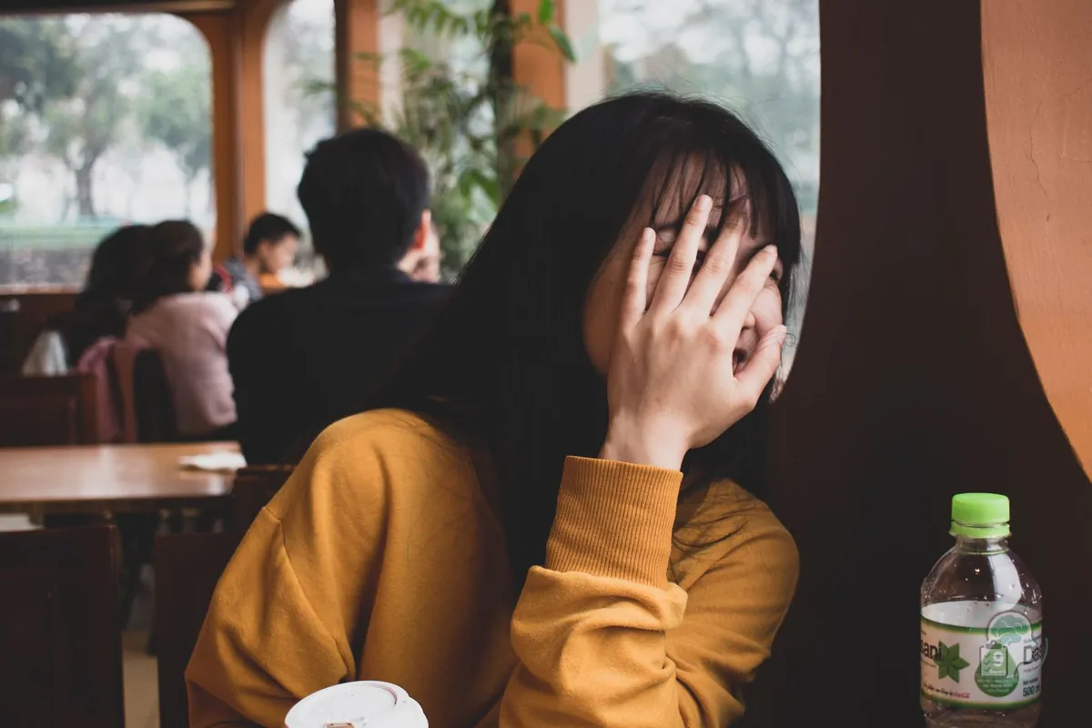 Joven en una cafetería que se tapa la cara con la mano entre risas, avergonzada, con otras personas sentadas detrás