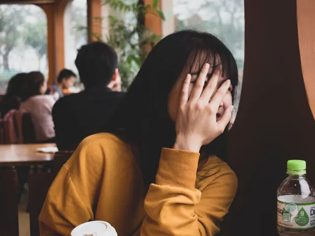 Joven en una cafetería que se tapa la cara con la mano entre risas, avergonzada, con otras personas sentadas detrás