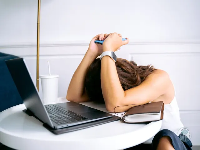 Mujer con la cabeza apoyada en la mesa junto al portátil y el móvil, bloqueada ante una tarea que no consigue empezar