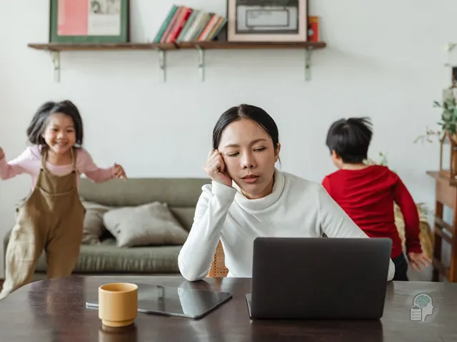 Madre agotada que apoya la cabeza en la mano delante del portátil mientras sus hijos corren por el salón, una imagen del burnout parental
