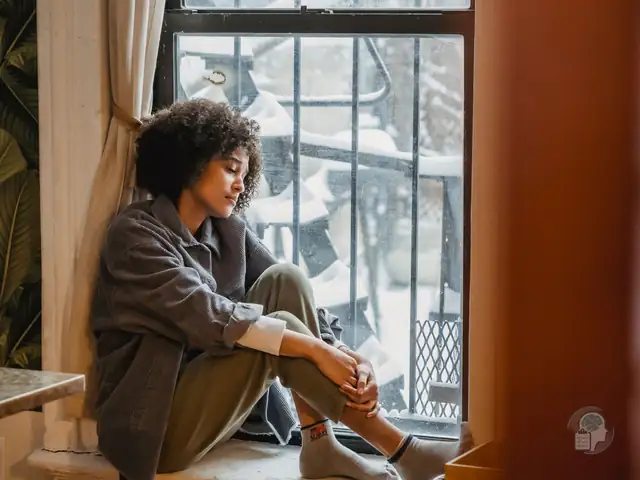 Mujer sentada en el alféizar abrazándose las rodillas junto a una ventana nevada, una imagen de la depresión estacional