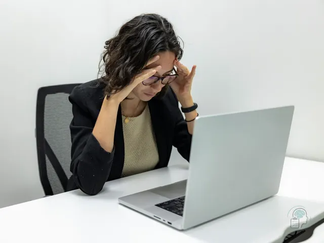 Mujer con gafas y americana negra sentada en su mesa de trabajo, con las manos en las sienes frente al portátil, una imagen del estrés laboral y el burnout