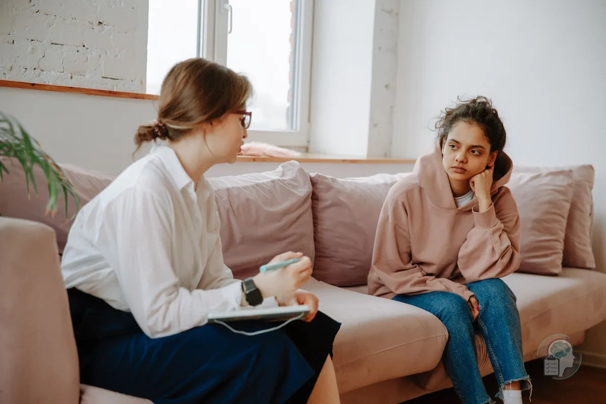 Psicóloga con gafas que habla con una adolescente de sudadera rosa sentadas en un sofá, durante una sesión de terapia para adolescentes