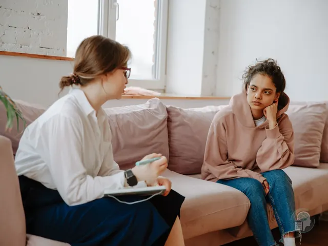 Psicóloga con gafas que habla con una adolescente de sudadera rosa sentadas en un sofá, durante una sesión de terapia para adolescentes