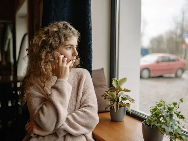 Mujer pensativa que habla por teléfono junto a la ventana de su casa, una escena habitual de la nostalgia de vivir en el extranjero