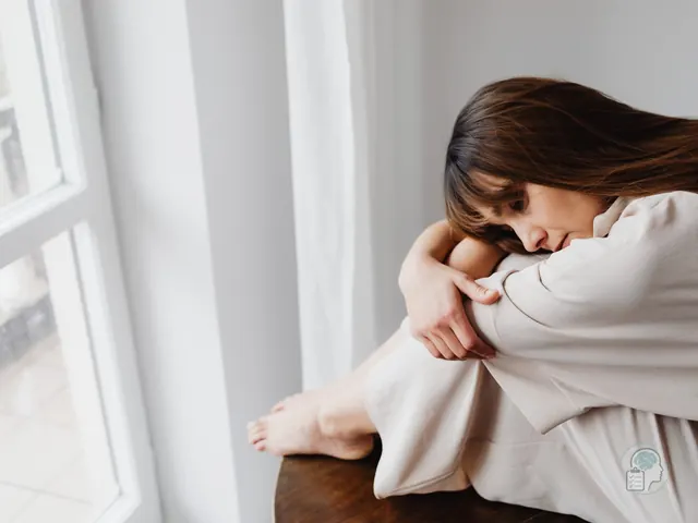 Mujer sentada junto a la ventana abrazándose las rodillas, desbordada por sus emociones