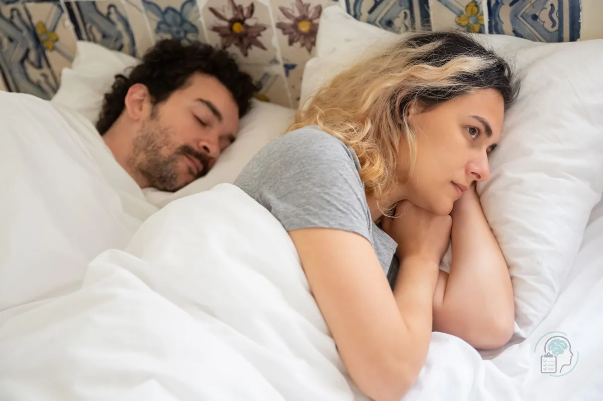 Mujer despierta en la cama de madrugada dándole vueltas a un pensamiento mientras su pareja duerme