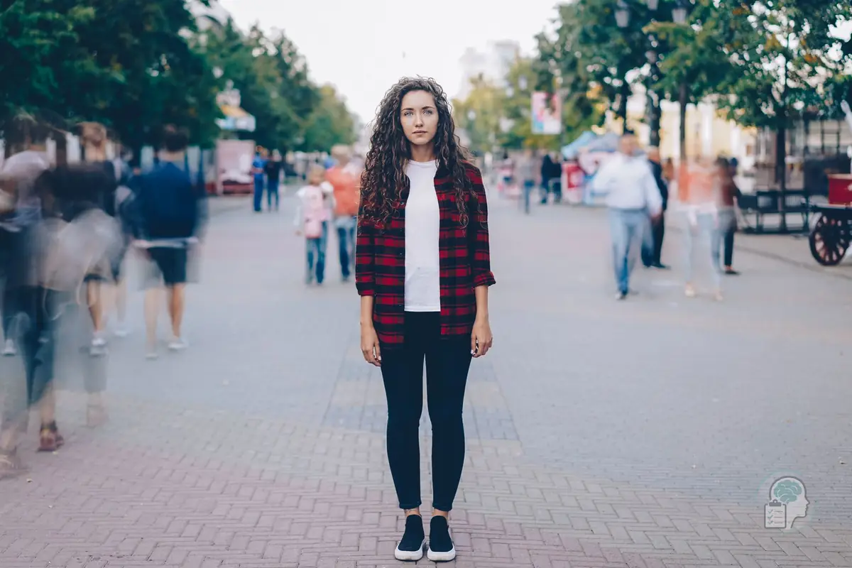 Mujer joven quieta en mitad de una calle peatonal mientras la gente pasa desenfocada a su alrededor, una imagen de sentirse diferente a los demás
