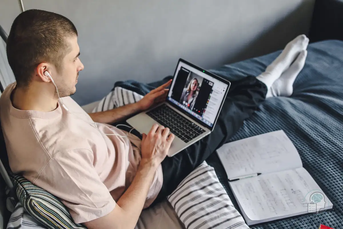 Joven con auriculares sentado en la cama durante una videollamada con el portátil, como en una sesión de terapia online para la ansiedad social