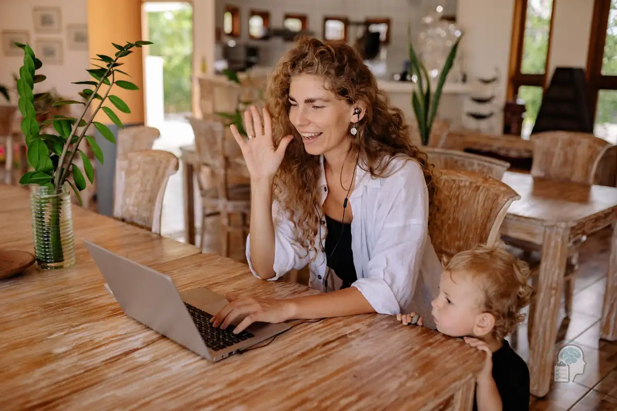Madre con auriculares que saluda en una videollamada con el portátil mientras su hijo pequeño mira a su lado, como en una sesión de terapia online desde casa