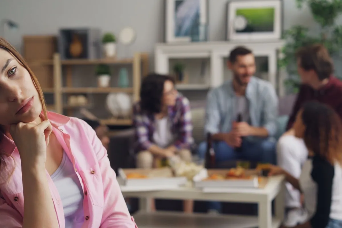 Mujer sola y pensativa en una fiesta mientras un grupo charla al fondo, ilustrando la ansiedad social que mide este test
