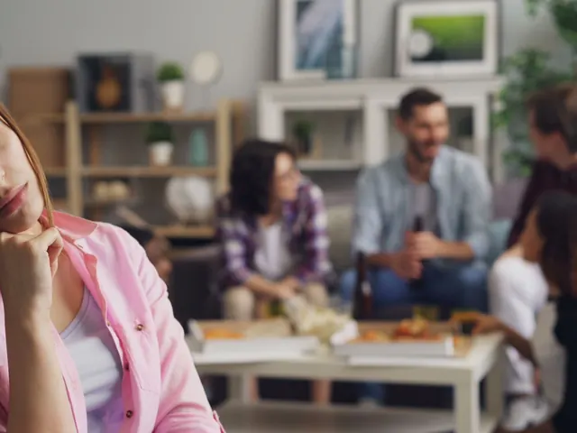 Mujer sola y pensativa en una fiesta mientras un grupo charla al fondo, ilustrando la ansiedad social que mide este test