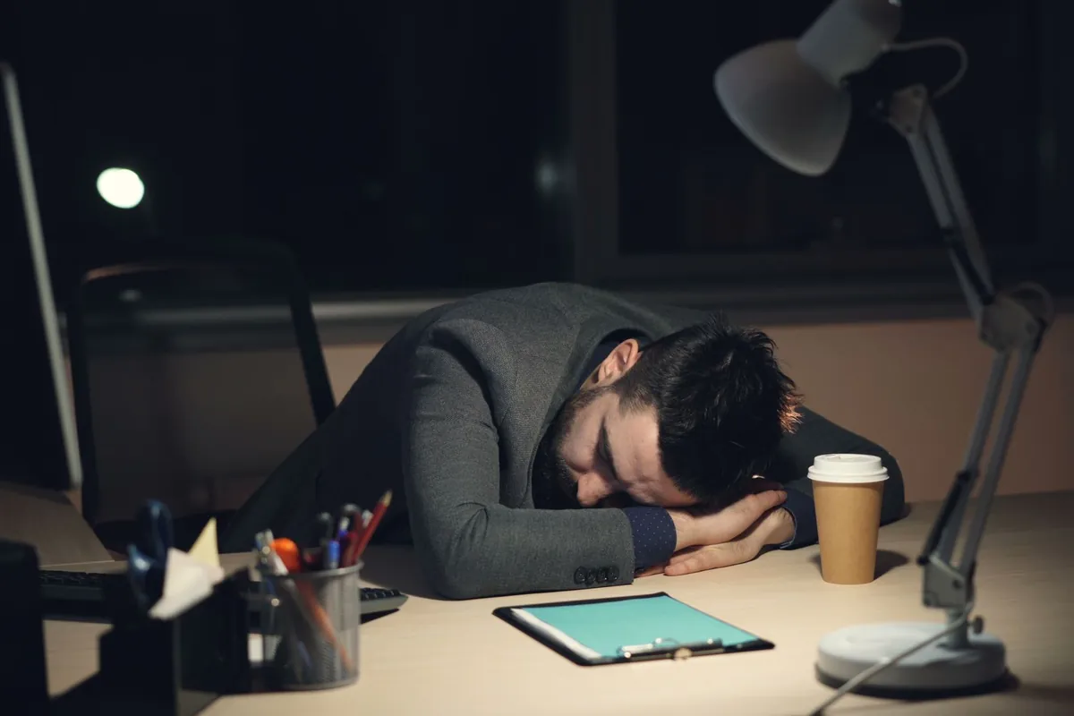 Persona dormida sobre el escritorio de noche con la lámpara encendida, imagen del agotamiento que mide este test de burnout