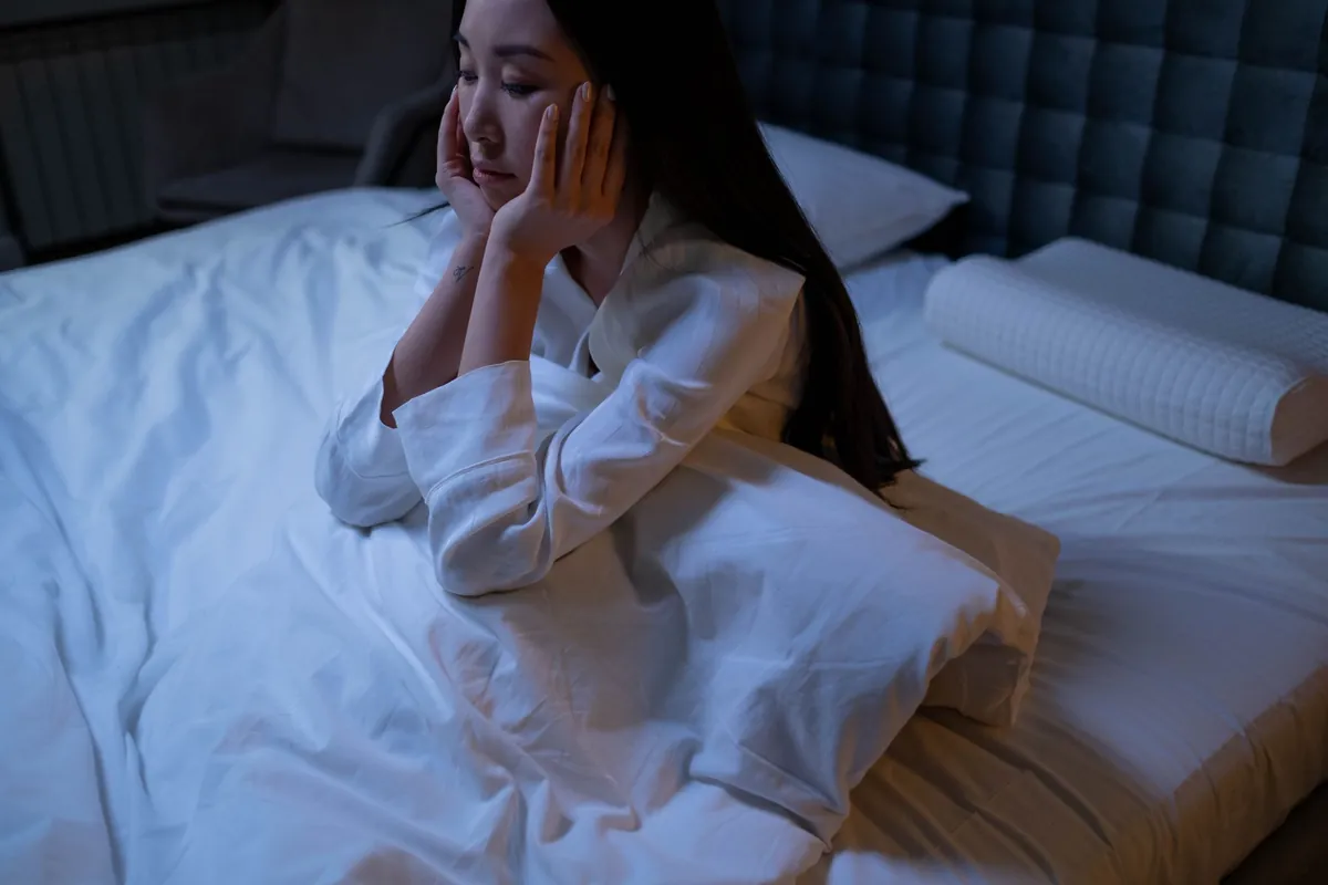 Mujer sentada en la cama de noche con las manos en la cara, sin poder dormir, ilustrando los síntomas del insomnio que valora este test